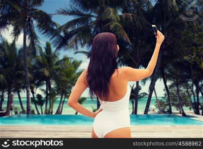 summer, travel, tourism, technology and people concept - sexy young woman taking selfie with smartphone over resort beach with palms and swimming pool background