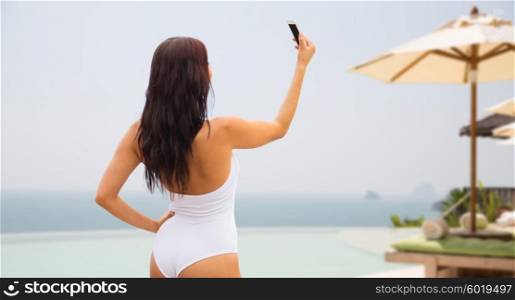 summer, travel, tourism, technology and people concept - sexy young woman taking selfie with smartphone over resort beach with parasols and swimming pool background