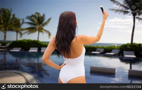 summer, travel, tourism, technology and people concept - sexy young woman taking selfie with smartphone over resort beach with palms and swimming pool background
