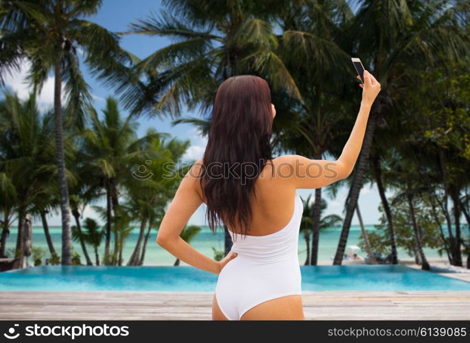 summer, travel, tourism, technology and people concept - sexy young woman taking selfie with smartphone over resort beach with palms and swimming pool background