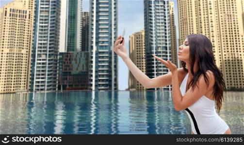 summer, travel, tourism, technology and people concept - sexy young woman taking selfie with smartphone and sending blow kiss over infinity edge swimming pool in dubai city background
