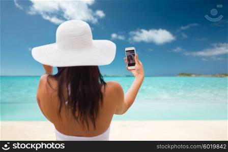 summer, travel, tourism, technology and people concept - smiling young woman or teenage girl in sun hat taking selfie with smartphone over tropical beach background