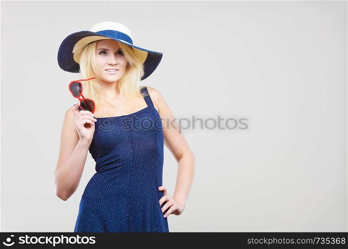 Summer trendy fashionable outfit ideas concept. Blonde attractive woman wearing short blue cocktail dress and sun hat, holding sunglasses, on grey. Woman wearing short navy dress and sun hat