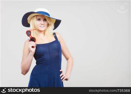 Summer trendy fashionable outfit ideas concept. Blonde attractive woman wearing short blue cocktail dress and sun hat, holding sunglasses, on grey. Woman wearing short navy dress and sun hat