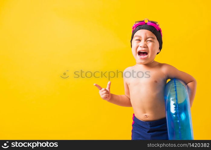 Summer vacation concept, Portrait Asian happy cute little child boy wear goggles and swimsuit hold blue inflatable ring, Kid hav fun point finger to side away, studio shot isolated yellow background