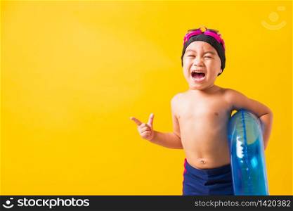 Summer vacation concept, Portrait Asian happy cute little child boy wear goggles and swimsuit hold blue inflatable ring, Kid hav fun point finger to side away, studio shot isolated yellow background