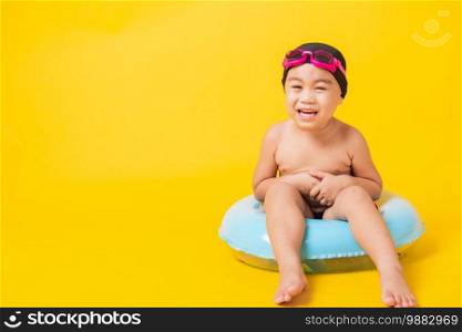 Summer vacation concept, Portrait Asian happy cute little child boy wear goggles, swimsuit hold beach blue inflatable ring, Kid have fun sit in inflatable, studio shot isolated yellow background
