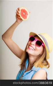 Summer vacation. Happy funny girl tourist in sunglasses and hat holding grapefruit citrus drinking juice from fruit on gray.