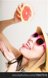 Summer vacation. Happy funny girl tourist in sunglasses and hat holding grapefruit citrus drinking juice from fruit on gray.