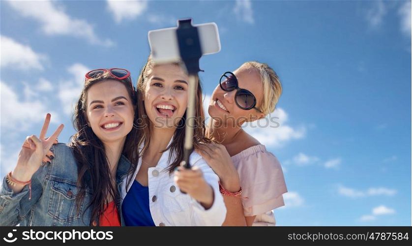 summer vacation, holidays, friendship, technology and people concept- group of smiling young women taking picture with smartphone on selfie stick over blue sky and clouds background