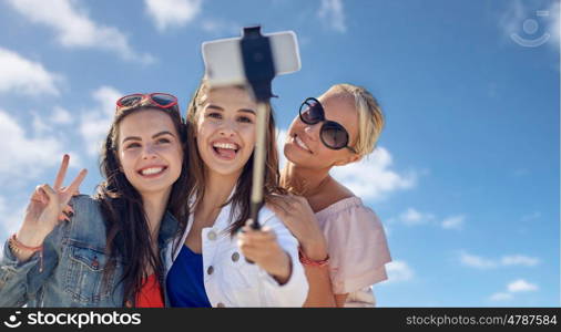 summer vacation, holidays, friendship, technology and people concept- group of smiling young women taking picture with smartphone on selfie stick over blue sky and clouds background