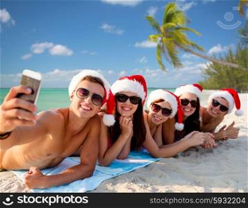 summer vacation, holidays, technology, travel and people concept - group of friends in santa helper hats taking selfie with smartphone on beach