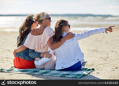 summer vacation, holidays, travel and people concept - group of smiling young women in sunglasses sitting on beach blanket and pointing finger to something