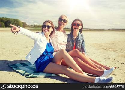 summer vacation, holidays, travel and people concept - group of smiling young women in sunglasses sitting on beach blanket and pointing finger to something. group of smiling women in sunglasses on beach