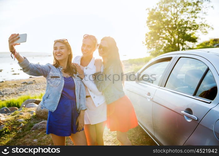 summer vacation, holidays, travel, road trip and people concept - happy teenage girls or young women with smartphone taking selfie near car at seaside