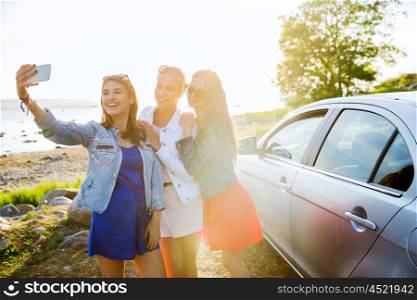 summer vacation, holidays, travel, road trip and people concept - happy teenage girls or young women with smartphone taking selfie near car at seaside