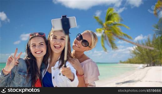 summer vacation, holidays, travel, technology and people concept- group of smiling young women taking picture with smartphone on selfie stick over tropical beach background