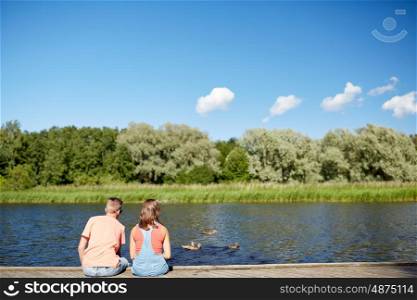 summer, vacation, love and people concept - happy teenage couple sitting on river berth looking at swimming ducks