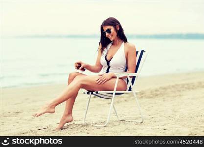 summer vacation, tourism, travel, holidays and people concept - close up of young woman in swimsuit sunbathing on folding chair and spraying sunscreen oil to her skin