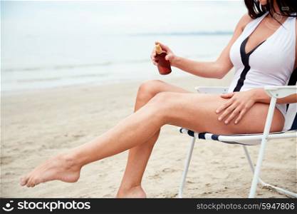 summer vacation, tourism, travel, holidays and people concept - close up of young woman in swimsuit sunbathing on folding chair and spraying sunscreen oil to her skin