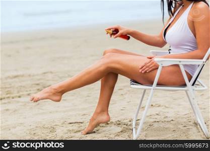 summer vacation, tourism, travel, holidays and people concept - close up of woman sunbathing in lounge and spraying sunscreen oil on beach