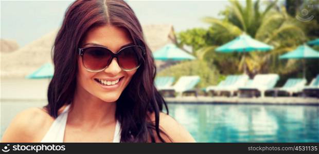 summer vacation, tourism, travel, holidays and people concept -face of smiling young woman with sunglasses over beach and swimming pool background