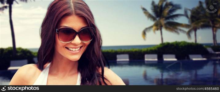 summer vacation, tourism, travel, holidays and people concept -face of smiling young woman with sunglasses over resort beach with palms and swimming pool background