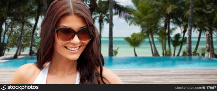 summer vacation, tourism, travel, holidays and people concept -face of smiling young woman with sunglasses over tropical beach with palms and swimming pool background