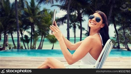summer vacation, tourism, travel, holidays and people concept - smiling young woman with tablet pc sunbathing in lounge or folding chair over tropical beach with palms and swimming pool background