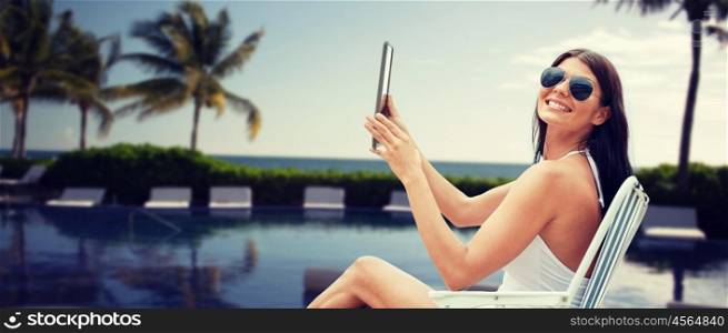 summer vacation, tourism, travel, holidays and people concept - smiling young woman with tablet pc sunbathing in lounge or folding chair over resort beach with palms and swimming pool background