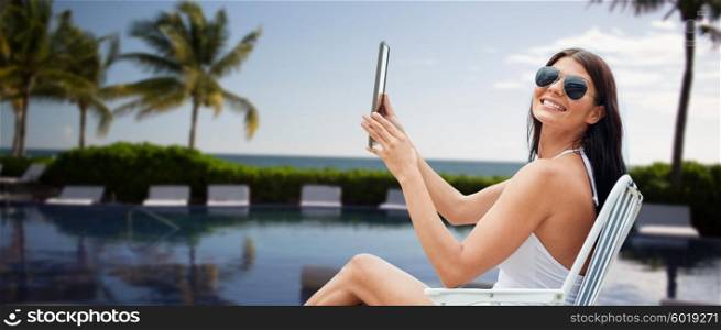 summer vacation, tourism, travel, holidays and people concept - smiling young woman with tablet pc sunbathing in lounge or folding chair over resort beach with palms and swimming pool background