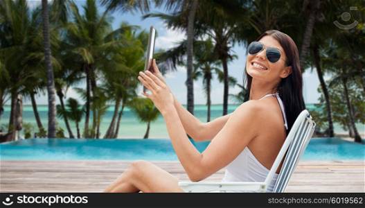 summer vacation, tourism, travel, holidays and people concept - smiling young woman with tablet pc sunbathing in lounge or folding chair over tropical beach with palms and swimming pool background