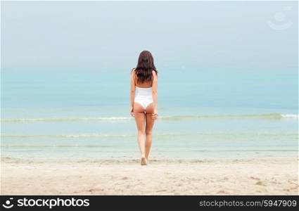 summer vacation, tourism, travel, holidays and people concept - young woman in swimsuit walking on beach from back
