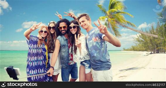 summer vacation, travel, technology and people concept - smiling young hippie friends in sunglasses taking picture by smartphone on selfie stick and showing peace gesture over beach background