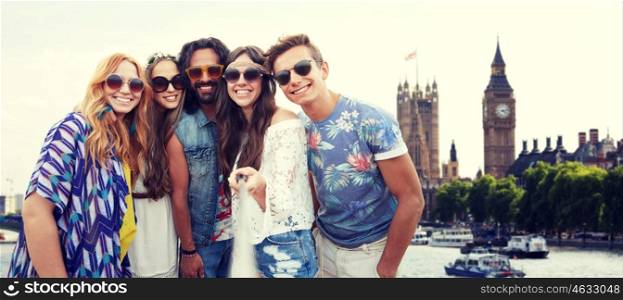 summer vacation, travel, tourism, technology and people concept - smiling young hippie friends taking picture by smartphone selfie stick over big ben tower in london city background