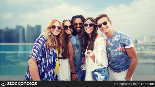 summer vacation, travel, tourism, technology and people concept - smiling young hippie friends taking picture by smartphone selfie stick over singapore city background