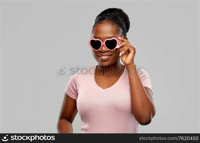 summer, valentine&rsquo;s day and eyewear concept - happy african american young woman in heart-shaped sunglasses over grey background. african american woman in heart-shaped sunglasses