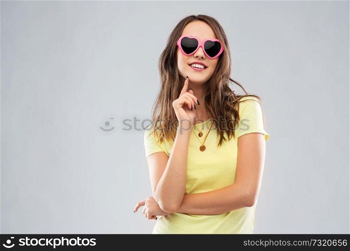 summer, valentine&rsquo;s day and people concept - smiling young woman or teenage girl in yellow t-shirt and heart-shaped sunglasses over grey background. teenage girl in heart-shaped sunglasses