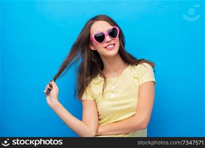 summer, valentine&rsquo;s day and people concept - smiling young woman or teenage girl in yellow t-shirt and heart-shaped sunglasses over bright blue background. teenage girl in heart-shaped sunglasses