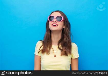 summer, valentine’s day and people concept - smiling young woman or teenage girl in yellow t-shirt and heart-shaped sunglasses over bright blue background. teenage girl in heart-shaped sunglasses