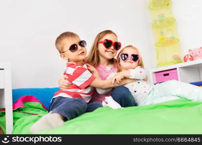 summer, valentines day and childhood concept - happy little kids in heart shaped sunglasses hugging at home. happy little kids in sunglasses hugging at home
