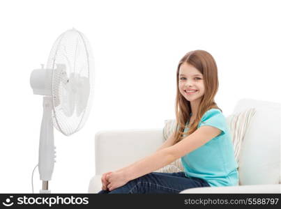 summer, weather and equipment concept - smiling little girl sitting on sofa with big fan at home