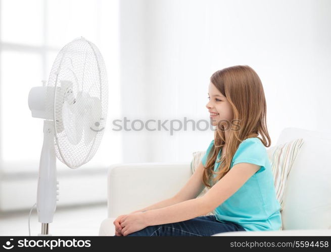 summer, weather and equipment concept - smiling little girl sitting on sofa with big fan at home