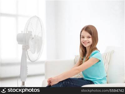 summer, weather and equipment concept - smiling little girl sitting on sofa with big fan at home