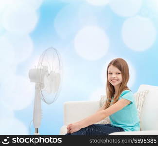 summer, weather and equipment concept - smiling little girl sitting on sofa with big fan at home