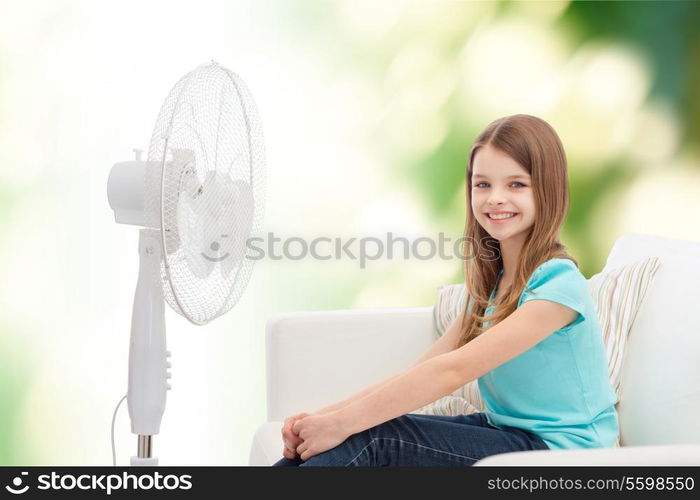 summer, weather and equipment concept - smiling little girl sitting on sofa with big fan at home