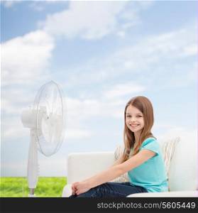 summer, weather and equipment concept - smiling little girl sitting on sofa with big fan at home