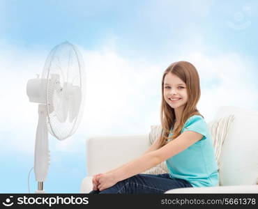 summer, weather and equipment concept - smiling little girl sitting on sofa with big fan at home