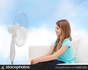 summer, weather and equipment concept - smiling little girl sitting on sofa with big fan at home