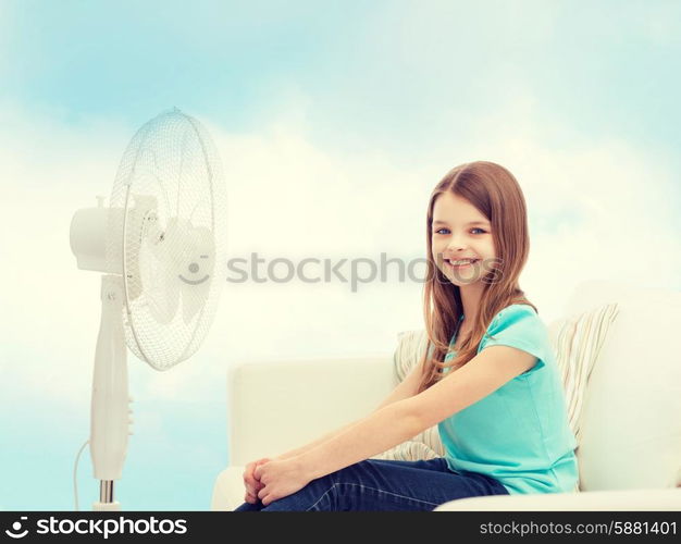 summer, weather and equipment concept - smiling little girl sitting on sofa with big fan at home